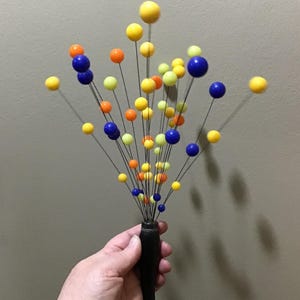 May include: A decorative arrangement featuring a black vase holding numerous stems, each topped with a colorful sphere. The spheres are in shades of yellow, orange, and blue. The arrangement is held in a hand.