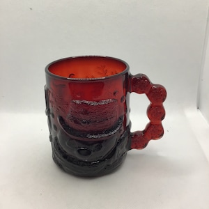 May include: A red glass mug with a textured design and a handle with a series of raised circles. The mug has a dark red color and a glossy finish.