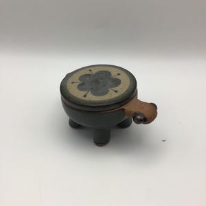 May include: A small, round, dark brown ceramic lidded container with three short legs. The lid features a light beige center with a dark gray, six-petaled flower design. A small, protruding handle extends from one side of the container.
