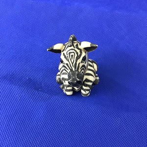 May include: A small, ceramic zebra figurine with black and white stripes. The zebra is in a seated position with its ears perked up. The figurine is on a blue surface.