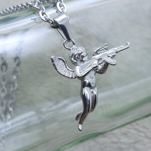 Pu&ograve; includere: Collana con catena d'argento con un ciondolo a forma di cherubino che tiene una pistola. Il cherubino ha le ali ed &egrave; in una posa di volo.