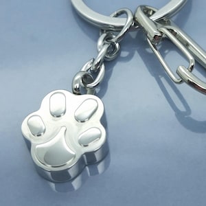 Puede incluir: Un llavero plateado con un dije de huella de pata. El dije es un diseño tridimensional con bordes redondeados y huellas de almohadillas detalladas. El llavero incluye una anilla partida y un mosquetón.