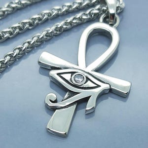 Puede incluir: Un collar con colgante plateado que presenta un símbolo Ankh con un diseño de Ojo de Horus. El colgante tiene un acabado pulido y una pequeña gema transparente en el ojo. La cadena es de estilo grueso y tejido.
