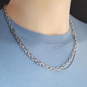 May include: A silver necklace featuring three different chain designs. The necklace is layered, with a curb chain, a box chain, and a chain with oval links. The necklace is shown against a light blue backdrop.