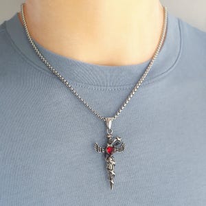 Può includere: Collana con pendente color argento con un design a pugnale e una pietra rossa a forma di cuore. Il pendente presenta ali e un serpente. La collana è indossata da una persona con una maglietta azzurra.