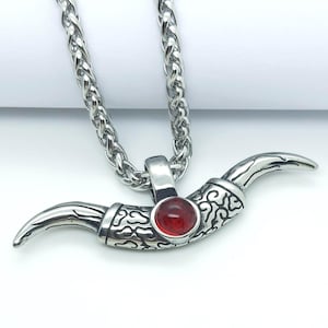 May include: A silver chain necklace with a silver pendant shaped like a bull's horn with a red gemstone in the centre.