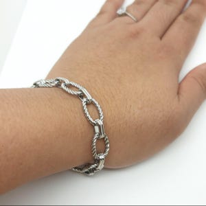 May include: A silver bracelet featuring oval links with a twisted rope design. The metal bracelet is worn on a wrist. The links are connected by a solid bar. The bracelet is a piece of jewellery.