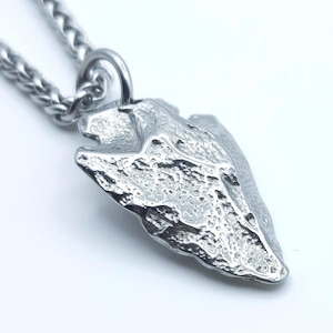 May include: A silver pendant necklace with a textured arrowhead design. The pendant is attached to a chain with a woven pattern. The arrowhead has a rugged, stone-like appearance, and the metal has a bright, reflective finish.
