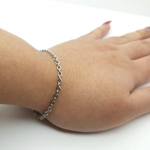 May include: A silver chain bracelet with an oval-link design. The bracelet is worn on a wrist, highlighting its simple and elegant style. The jewellery is made of a shiny, metallic material.