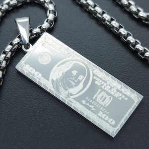 May include: A silver-colored pendant necklace featuring a rectangular pendant engraved with a $100 bill design. The word "SUPREME" is at the top. The chain is a box link style.