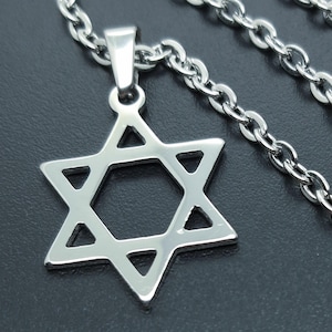 Puede incluir: Un collar con colgante de estrella de David plateado. El colgante es una estrella de seis puntas con un centro hueco, suspendida de una cadena a juego. La cadena tiene un diseño de eslabones. El collar se muestra sobre un fondo oscuro.