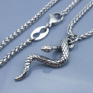 May include: A silver-toned metal necklace featuring a detailed snake pendant with a textured scale pattern. The pendant is attached to a wheat chain that includes a lobster claw clasp and an oval-shaped link near the closure mechanism.
