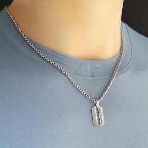 Könnte beinhalten: Silberfarbene Halskette mit einem Anhänger in Form einer Rasierklinge. Der Anhänger hängt an einer Weizenkette. Die Person im Bild trägt ein hellblaues T-Shirt.