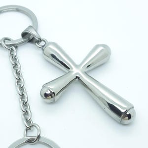 Op de afbeelding: Een zilverkleurige kruisvormige hanger sleutelhanger. Het kruis heeft afgeronde randen en een gepolijste afwerking. De sleutelhanger bevat een ketting en een ring voor het bevestigen van sleutels.