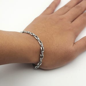 Puede incluir: Una pulsera de cadena de metal en tono plateado que presenta una serie de eslabones alargados, lisos y pulidos, conectados por pequeños espaciadores circulares redondeados, creando una banda continua, flexible y reflectante.