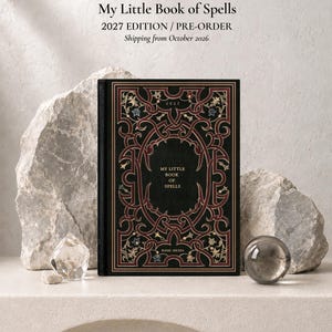 Op de afbeelding: Een zwart hardcover boek getiteld My Little Book of Spells met ingewikkelde rode en gouden filigraanpatronen op de kaft. De tekst 2027 en Basic Hexes staat op de voorkant gedrukt. Het boek staat tussen twee grote, grillige grijze rotsen in.