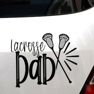 May include: Black vinyl decal with the words "Lacrosse Dad" in a bold font, with two crossed lacrosse sticks and radiating lines. The decal is designed to be applied to a car or other surface.