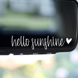 May include: A white vinyl decal applied to a black rearview mirror surface. The decal features the text "hello sunshine" in a cursive script font, followed by a small, solid white heart icon positioned to the right of the text.