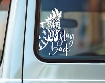 May include: A white vinyl decal applied to a vehicle window featuring a stylized graphic of a chicken head with a stern expression, accompanied by the text "stay back" written in a flowing, cursive script font below the bird illustration.