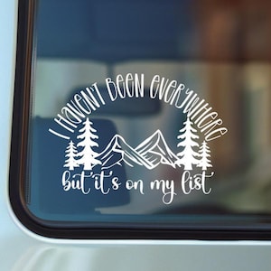 May include: White decal with the text "I haven't been everywhere but it's on my list." The design includes mountains and pine trees. The decal is likely for a car window or other smooth surface.