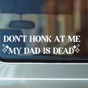 May include: White vinyl car decal with the text "DON'T HONK AT ME MY DAD IS DEAD" in a bold, sans-serif font. The decal includes star and diamond shapes on either side of the text. The decal is applied to a car window.