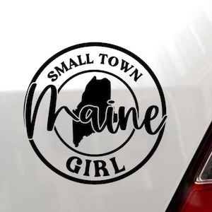 May include: A black circular vinyl decal featuring the text SMALL TOWN GIRL in a bold sans-serif font. In the center, a solid black silhouette of the state of Maine is overlaid with the word Maine written in a large black script font.