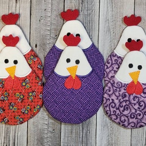 May include: Set of three quilted hot pads shaped like chickens. The hot pads are red with white floral print, purple with a geometric pattern, and purple with a swirl pattern. Each hot pad has a white chicken head with red comb and beak.