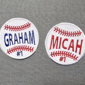 May include: Two white baseball-themed patches with red stitching. The top patch says "GRAHAM #1" in blue lettering. The bottom patch says "MICAH #1" in red lettering.