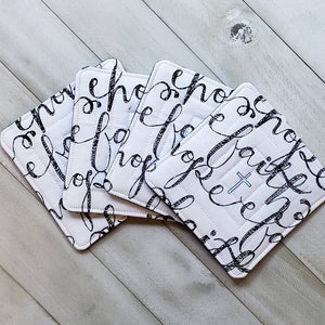 May include: Set of four white fabric coasters with black cursive writing that says "Hope, Faith, Love" and a small cross.