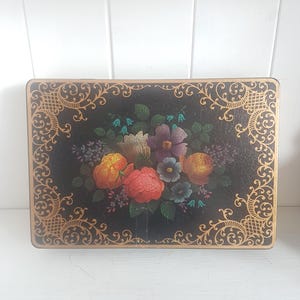 Könnte beinhalten: Eine antike Holzkiste mit einer schwarz lackierten Oberfläche und einem goldenen Blumendesign. Die Kiste zeigt ein gemaltes Blumenbouquet mit roten, gelben, orangefarbenen, violetten und blauen Blumen.