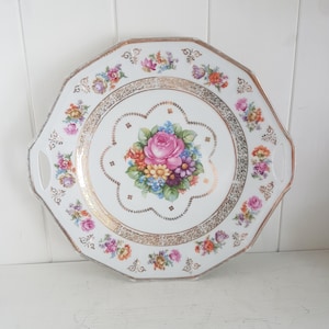 May include: White porcelain serving tray with a gold rim and handles. The tray features a central floral bouquet with a pink rose and colourful blossoms. Smaller floral arrangements and gold accents decorate the border.