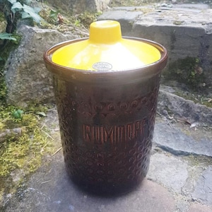 Op de afbeelding: Een bruine keramische pot met een geel deksel. De pot heeft een getextureerd oppervlak en het woord "RUMTOPF" is erop gegraveerd.