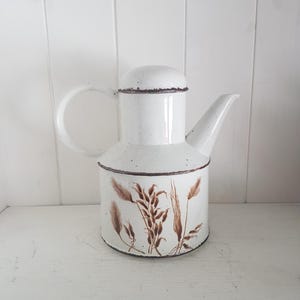 Könnte beinhalten: Weißer Emaille-Krug mit gebogenem Griff und Ausguss. Der Krug hat ein braunes Weizenähren-Design um den Sockel und eine passende braune Zierleiste. Der Krug steht auf einer weißen Oberfläche.