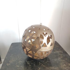 Può includere: Un oggetto decorativo sferico color oro con intricati motivi traforati. La sfera ha un piccolo anello nella parte superiore per appenderla. L'oggetto è appoggiato su una superficie scura e usurata.