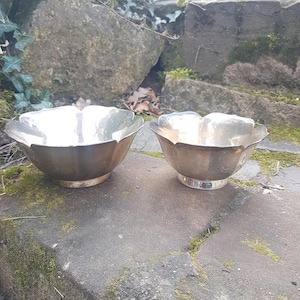 Pode incluir: Duas tigelas de metal prateado com uma borda em forma de pétala. As tigelas estão sobre uma superfície de pedra. As tigelas têm uma base redonda e um acabamento brilhante. As tigelas são provavelmente para fins decorativos ou de serviço.