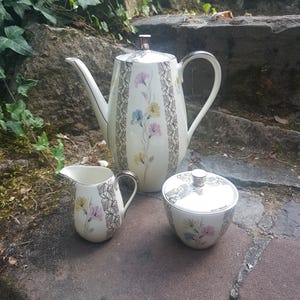 Può includere: Un servizio da tè vintage color crema con una teiera alta, una lattiera e una zuccheriera. Il set è decorato con motivi floreali rosa, gialli e blu e rifinito con un design in pizzo grigio e bianco. La teiera ha un manico e un coperchio argentati.