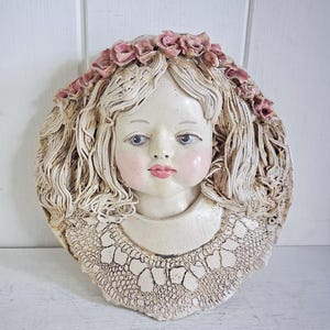 May include: A circular ceramic wall plaque featuring a sculpted face with wavy hair adorned with a crown of pink roses. The figure has blue eyes, pink cheeks, and pink lips, wearing a textured collar with a lace-like pattern in an off-white finish.