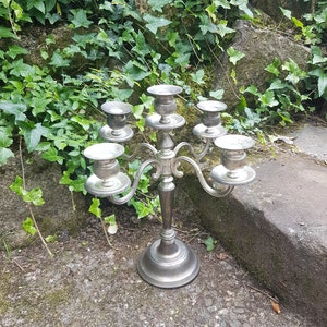 May include: Silver candelabra with five candle holders, featuring a decorative, ornate design. The candelabra is standing on a gray, pebbled surface.