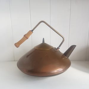 Könnte beinhalten: Ein kupferfarbener Wasserkocher mit Holzgriff und Ausguss. Der Griff besteht aus hellem Holz und ist mit einem Metallrahmen am Wasserkocher befestigt. Der Wasserkocher hat einen abgerundeten Körper und einen kleinen Deckel.