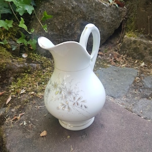 Könnte beinhalten: Weißer Keramikkrug mit bauchigem Körper, gebogenem Henkel und Ausguss. Der Krug ist mit einem floralen Muster in Gold und Grau verziert. Der Krug steht auf einer Steinoberfläche.