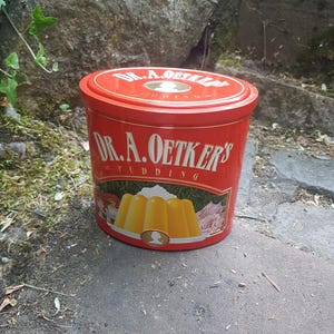 Könnte beinhalten: Eine rote Dr. A. Oetker's Puddingdose mit Deckel. Die Dose zeigt den Markennamen und ein Bild von gelbem Pudding mit Schlagsahne und Früchten. Die Dose ist zylindrisch und steht auf einer Steinoberfläche.