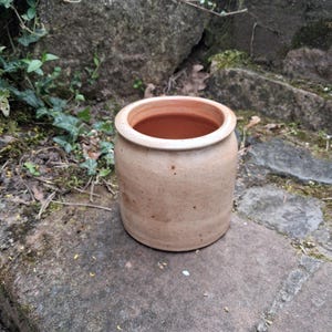 May include: A handmade, cylindrical, beige ceramic pot with a wide opening. The pot has a slightly textured surface and is sitting on a stone surface. The interior of the pot is a darker brown color. The pot is likely used for storage or decoration.