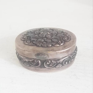 Könnte beinhalten: Kleine, runde, antik-silberfarbene Schatulle. Der Deckel hat ein Blumenmuster, und die Basis hat verzierte Schnörkel. Die Schatulle scheint aus Metall zu sein und hat ein angelaufenes Aussehen, was auf das Alter hindeutet. Ein dekorativer Artikel.