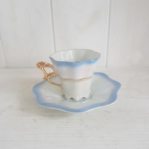 Op de afbeelding: Een vintage theekop en schotel set. De kop en schotel zijn wit met een lichtblauwe rand. De kop heeft een gouden oor en een gouden band. De schotel heeft een geschulpte rand.