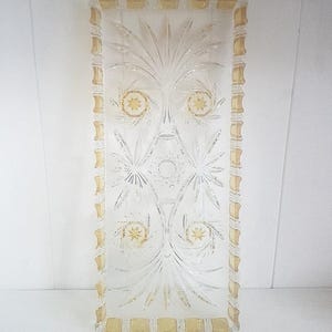 Puede incluir: Bandeja rectangular de vidrio transparente con intrincados diseños florales y de estrellas grabados. La bandeja tiene un borde dorado con un patrón rectangular repetitivo. El diseño presenta patrones simétricos y una estrella central.