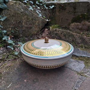 Op de afbeelding: Een ronde keramische kom met deksel, crème van kleur. Het deksel heeft gouden en groene geometrische patronen. Een dunne turquoise streep accentueert de rand. De kom heeft een diameter van ongeveer 20 cm.