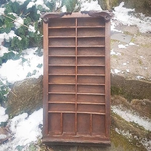 May include: A vintage wooden display cabinet with multiple small compartments. The cabinet is rectangular with a dark brown finish and decorative carvings at the top. The cabinet is empty and appears to be mounted on a stone wall with snow and greenery in the background.