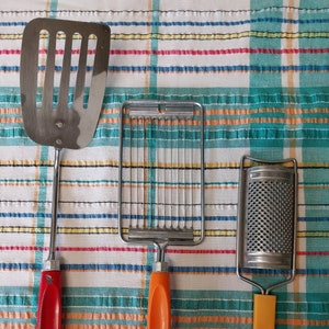 May include: Three kitchen utensils with colorful handles: a red handled spatula, an orange handled waffle cutter, and a yellow handled grater.