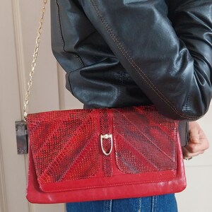 May include: A red clutch bag with a gold chain strap. The bag has a textured, snake-like pattern on the flap and a gold-coloured clasp. The bag is held against a person wearing a black leather jacket and blue jeans.