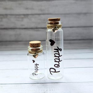 May include: Two clear glass bottles with cork stoppers. The bottles are decorated with twine, small heart charms, and the word "Poppy" in black script. One bottle is taller than the other.